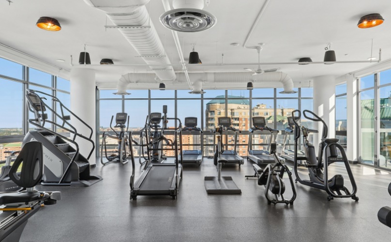 Fitness center with cardio and weights surrounded by floor-to-ceiling windows  At J Sol Apartments In Arlington, VA